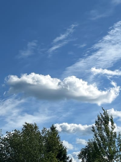 The image depicts a clear blue sky with scattered white clouds. In the foreground, the tops of green trees are visible, suggesting an outdoor setting in a location with vegetation. The lighting indicates it is daytime. The provided location information, "Слава Снт, Russia," specifies a settlement named "Slava SNT" in Russia. The scene is natural and tranquil, with no human presence or specific urban features evident in the frame.