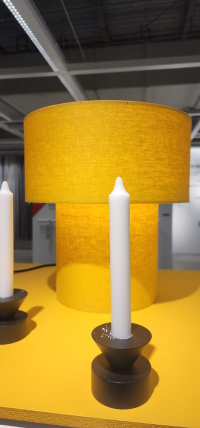A close-up, eye-level shot captures a bright yellow, cylindrical table lamp with a textured fabric shade. Two white taper candles are positioned in front of the lamp. The candle on the right is slightly closer to the camera and is centrally framed. The candle on the left is partially visible and positioned further back. Both candles are placed in black, cone-shaped candle holders with a cylindrical base. The objects rest on a flat, yellow surface that extends to the bottom of the frame. In the background, the ceiling is visible with exposed lighting fixtures and structural elements, suggesting an indoor setting, likely a store or exhibition space in Gwangmyeong, South Korea. The lighting is bright and even, illuminating the subjects and creating soft shadows.