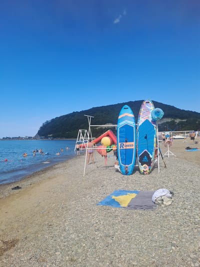 A wide, pebbled beach in Dzhubga, Russia, slopes down to a calm, blue sea under a clear sky. Several people are visible in the water, some on inflatable rings, enjoying the refreshing swim. On the beach, two vibrant paddleboards stand upright, leaning against a white metal frame. One paddleboard is predominantly blue with an orange trim and "RUneed" written vertically. The other is a colorful, patterned board. Nearby, a yellow inflatable structure and a red inflatable slide suggest a children's play area. A grey towel and some clothing are laid out on a map-patterned blanket on the pebbles, with a beige backpack resting on top. The shoreline is lined with a thin strip of sand, with scattered debris and a single black sandal. In the background, a lush, tree-covered mountain range forms a dramatic backdrop to the seaside activity. Buildings and structures are visible along the coastline in the distance.