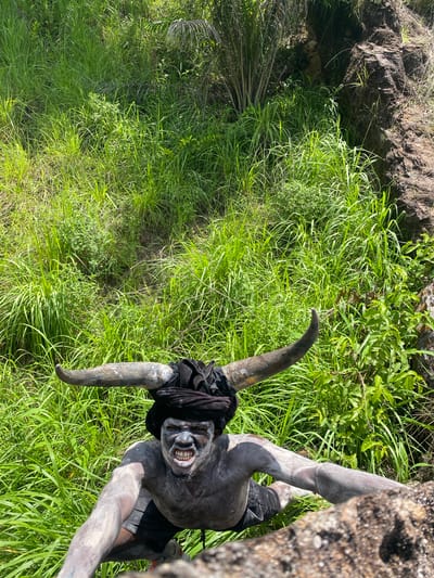 The image shows a person costumed as a horned figure with gray body paint in a grassy outdoor area. The figure has prominent horns, a dark garment draped over their head and torso, and black shorts. The person is positioned against a rock or tree trunk. The background features lush green grass and vegetation. The location is not discernible from the image.