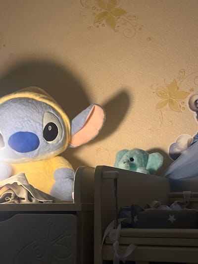 The image shows a stuffed toy Stitch with a yellow hoodie, a teal elephant plush toy, and part of a blue stuffed toy. The toys are placed on a shelf or furniture. The background is a wall with a textured wallpaper featuring floral patterns. The lighting suggests an indoor setting, possibly a bedroom, with a shadow cast from the Stitch toy. The location is identified as Pikalyovo, Russia.