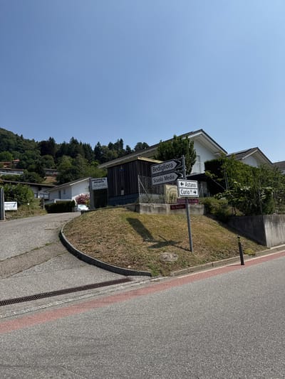 This is an exterior, eye-level, medium shot of a street scene in Lema, Switzerland, on a sunny day. The road curves from the left foreground to the right. In the mid-ground, a multi-directional street signpost stands on a grassy slope. The signpost has four signs. The top sign reads "Bedigliora" with an arrow pointing left. Below that, a sign reads "Scuola Media" with an arrow pointing left. To the right of these, a sign reads "Astano" with an arrow pointing left, and below that "Curio" with an arrow pointing right. At the base of the signpost, there are two smaller signs on a horizontal bar, one reading "Via Crussetta" and the other "Via Banghes."
Behind the signpost, a low concrete retaining wall is visible, topped with a fence and dry grass. Beyond the fence, there is a wooden building with a dark roof. Further back, a white building with a tiled roof is partially obscured by trees and bushes. To the far left of the image, a white sign with dark text is partially visible, and a dark grate runs along the edge of the road in the left foreground. The background is dominated by a lush green hillside covered in trees. The sky is clear and blue.