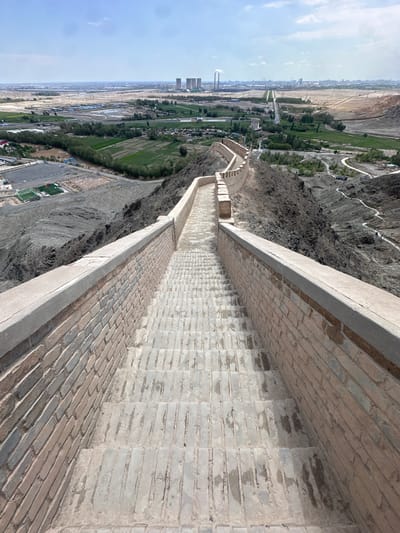 The image shows a stone staircase descending along a section of the Great Wall in Jiayuguan, China. The staircase is constructed from weathered bricks and is flanked by a stone wall. Beyond the wall, the landscape features rugged, arid terrain with some sparse vegetation and a winding path. In the distance, the cityscape of Jiayuguan is visible, characterized by industrial structures, including cooling towers of a power plant, and agricultural fields. The sky is bright and partly cloudy.