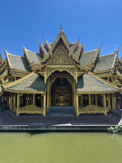 The image shows a detailed view of a traditional Thai pavilion located within the Ancient City (Muang Boran) in the Bang Pu Subdistrict Municipality, Thailand. The pavilion has a tiered roof design with green tiles, ornate gold trim, and intricate carvings. A golden Buddha statue is visible inside the pavilion. The pavilion is situated next to a body of water. The sky is clear and blue.