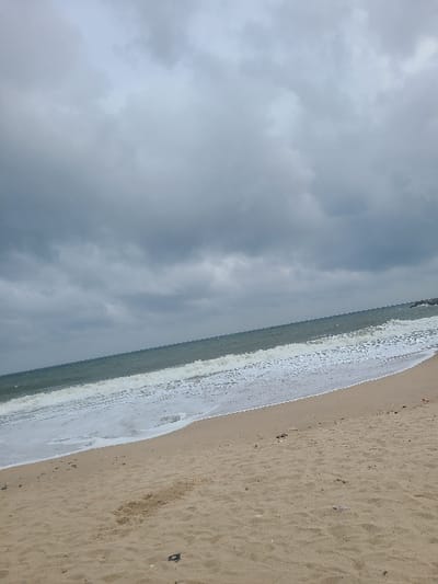 The image displays an expansive coastal scene, identified as a beach in Ibeju, Nigeria, under an overcast sky. The upper portion of the frame is dominated by a dense formation of grey clouds, suggesting heavy atmospheric conditions. Below the horizon line, a body of water, likely the Atlantic Ocean, extends with dark green to teal coloration. Multiple waves are observed breaking onto the shore, generating significant white foam as they recede. The lower half of the image consists of a light tan sandy beach, exhibiting variations in texture and moisture content. Scattered dark specks, possibly small debris, are visible on the sand, with a more distinct dark, irregularly shaped object present in the immediate lower-left foreground. A faint, dark structure, consistent with either a rocky outcrop or a constructed barrier, is discernible at the far right edge where the water meets the distant land. No individuals, animate subjects, or explicit actions are depicted within the scene.