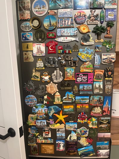 The image is a close-up, eye-level, full shot of a refrigerator door covered in numerous novelty magnets, predominantly from various travel destinations. The magnets are a mix of rectangular, circular, and irregularly shaped items, featuring landmarks, cityscapes, flags, and cultural symbols. Some magnets depict famous structures like the Eiffel Tower, the Leaning Tower of Pisa, the Burj Khalifa, and various cathedrals and mosques. Other magnets show stylized representations of animals, food items, and maps. The colors of the magnets range from vibrant hues to muted tones, and many have embossed or raised details. The magnets are arranged in a dense, overlapping pattern across the dark gray surface of the refrigerator door. There are no people visible in the image. The scene appears to be indoors, with the lighting suggesting it is an ambient light source. The lower left portion of the image shows a partial view of a white door with a black door handle. The context does not provide any information that would place this scene in Novaya Adygeya, Russia; all identifiable locations are from diverse international destinations.
