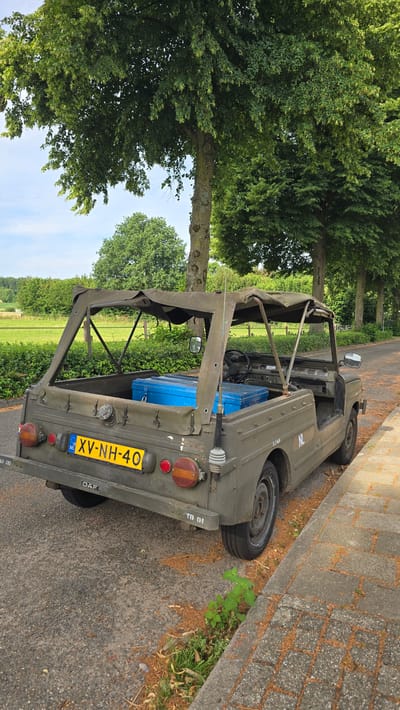 A military-style vehicle, possibly a DAF, is parked on the side of a paved road in Zeddam, Netherlands. The vehicle is olive green and has a canvas top. Two bright blue storage containers are visible in the open cargo area of the vehicle. The license plate reads "XV-NH-40". The road is lined with trees and green grass. The sky is blue with some clouds. The scene appears to be a sunny day.