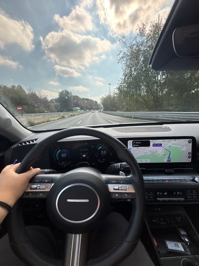 An interior view from a vehicle in motion captures a multi-lane asphalt highway under a partly cloudy sky near Cardano al Campo, Italy. The driver's left hand, wearing a black wristband, holds the black and chrome steering wheel of a modern car. The digital instrument cluster displays a speed of 85 km/h and an "ECO" mode with a value of "2.0". To the right, an infotainment screen shows a navigation map indicating the current location in the area of Cardano al Campo, Italy, and a destination 9.3 km to Milan. Below the screen, the center console features digital climate controls set to 21°C and a storage area containing small objects, including a QR code sticker. Through the windshield, minor dust is visible. The highway is flanked by metal guardrails on the right and dense green trees and vegetation on both sides. A circular "no entry" sign is discernible on the left side of the road in the distance. Several other vehicles are visible ahead, and bright sunlight causes glare from the upper right portion of the sky.