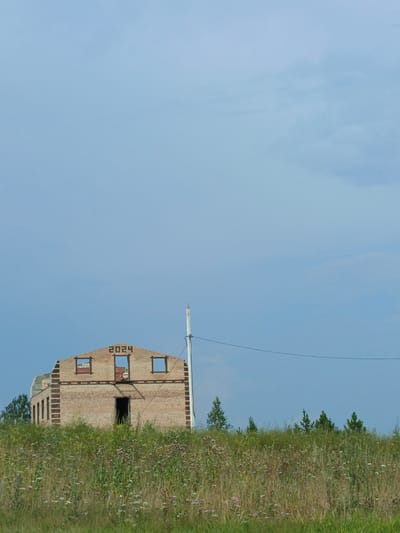 A partially constructed, two-story brick building with an unfinished gable roof stands in the foreground, against a bright, cloudy sky. The year "2024" is visible in dark brickwork on the upper gable. The building has open window frames and a dark doorway. A white pole, possibly a utility pole, stands to the right of the building, with a thin wire extending from it and across the frame. A grassy, overgrown field with wildflowers extends from the bottom of the image to the base of the building. Scattered trees are visible behind the building and to the left. The scene appears to be located in a rural or undeveloped area, possibly on the outskirts of Nizhnii Atlyan, Russia. There are no people visible in the image.