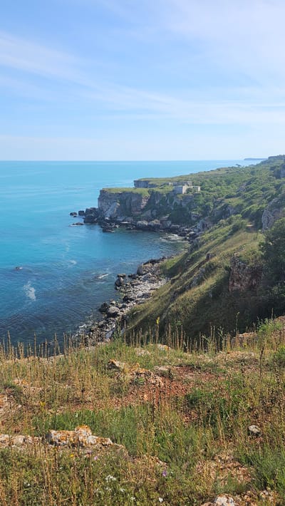 The image depicts a coastal scene in Kamen bryag, Bulgaria. A rocky cliff edge, covered with green and yellow grasses and low-lying vegetation, dominates the foreground. Below the cliff, the Black Sea appears in shades of turquoise and blue, with rocky outcroppings extending into the water. A few small boats are visible on the water. The sky is pale blue with faint clouds. In the distance, the cliff continues along the coastline. A small, white building sits atop the cliff.