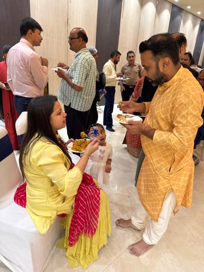 This media file captures an indoor social gathering taking place in New Town, India. Numerous individuals of various ages, including adults and at least one young child, are present, engaged in eating and socializing. The scene has a warm and communal atmosphere.

In the foreground, a woman wearing a yellow top and a red patterned skirt/dupatta is seen holding a plate of food, possibly rice or biryani, and appears to be feeding a young child dressed in a white frock. The child looks up with an open mouth, eagerly awaiting the food. To the right of the woman, a man in a yellow patterned kurta and white pants is also holding a plate with food, looking down at his plate or the food. His attire suggests traditional Indian wear.

Several other men are visible in the background, also holding plates and interacting. One man in a light plaid shirt is prominent on the left side, while another in a light pink shirt stands in the very foreground on the left. The attire of the attendees varies, with some wearing traditional Indian clothing like kurtas and others in more casual shirts.

The setting appears to be a large indoor hall or room with light-colored flooring and beige walls. The lighting is artificial, suggesting the event could be taking place at any time of day. No specific outdoor elements or weather conditions are visible. There is no discernible text in the image. The overall mood conveyed is one of a festive or celebratory meal, with people enjoying food and company.