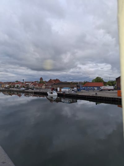 The image captures a scene in Køge, Denmark, featuring a canal reflecting the town's architecture and cloudy sky. On the left side of the canal, a row of buildings with red-tiled roofs lines the waterfront, interspersed with trees and vehicles. A pier with docked boats and a few parked trucks extends into the canal. The canal water mirrors the town and sky. The sky is overcast.