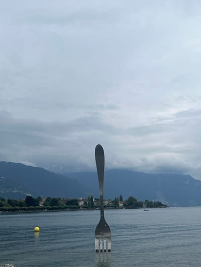 A large, vertical, polished metal fork sculpture, approximately 8 meters tall, emerges from the calm, dark blue-grey waters of Lake Geneva in Vevey, Switzerland. The sculpture, known as The Fork, is situated in the foreground, its four tines resting on the lakebed. To the left of the sculpture, a single bright yellow spherical buoy floats on the water's surface. In the midground, beyond the lake, a city shoreline with buildings and green trees is visible. The background is dominated by a series of large, dark mountains, their lower slopes partially obscured by a light haze or cloud cover. The sky is overcast and uniformly light grey, indicating a lack of direct sunlight. No individuals or dynamic actions are depicted in the static scene.