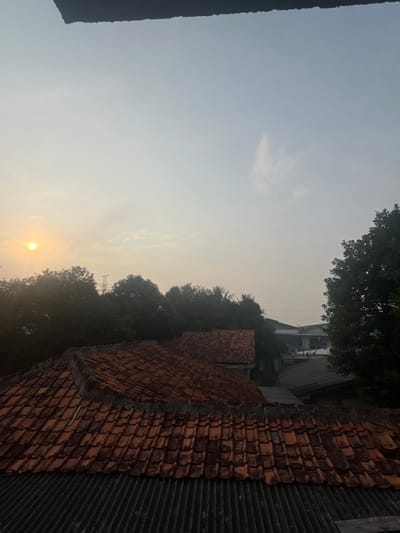 A hazy, overcast sky at sunrise partially obscures a sunlit disc on the left side of the frame. The foreground is dominated by a series of weathered, terracotta-tiled rooftops. Below these, corrugated metal sheeting is visible. Beyond the roofs, a dense line of dark green trees stretches across the middle ground, with a faint electrical tower visible behind them on the left. In the background, various buildings with darker roofs are scattered amongst the trees, suggesting a semi-urban or suburban environment. The overall scene depicts an early morning view over a residential area in Pasir Gadung, Indonesia.