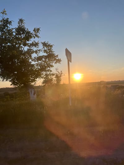 The image depicts a rural landscape at sunset, likely on the outskirts of Karagash, Moldova. The foreground consists of a dirt path and overgrown vegetation, with a tree on the left side of the frame. A signpost stands near the center, bearing a circular symbol with what appears to be numbers inside, possibly indicating a speed limit or a zone restriction. The sun is low on the horizon, casting a warm, golden light across the scene and creating lens flare. In the background, a hilly terrain is visible with scattered structures and trees. The sky is a clear blue transitioning to orange and yellow hues near the horizon, indicating the end of the day. There are no people or animals visible in the image.