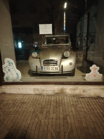An old silver Citroen 2CV car sits in a storefront window. The license plate reads "6155 ZC 84." A white sign is visible above the car. On either side of the car, cut-out shapes of a tower and a fountain are placed against the glass. The building's exterior has a brick pavement leading up to the window, with a white stone base. Lighting appears dim, suggesting it is nighttime.