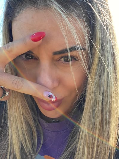 A close-up, head-and-shoulders selfie shows a woman with blonde hair and makeup. She is holding her right hand up, covering her left eye and part of her forehead. Her fingernails are decorated with red polish and white dots on the index finger, and on her middle finger, a pink nail has a ladybug and flowers painted on it. A ring with a diamond is visible on her ring finger. A rainbow-colored light flare cuts across the lower part of the image. She is wearing a purple t-shirt and a dark necklace. The background appears to be an outdoor setting with sunlight. The location of Ravnogor, Bulgaria is not determinable from the image.