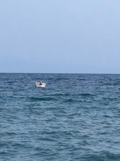 A small white boat with a console and seats is floating on the calm, deep blue waters of the sea. The sky above is a pale, hazy blue, suggesting either early morning or late evening. The horizon is clearly defined where the water meets the sky. No people are visible on the boat or in the surrounding water. The scene is tranquil and minimalist, with the boat being the sole focal point against the vast expanse of sea and sky. This photograph could have been taken from the shore or another vessel.