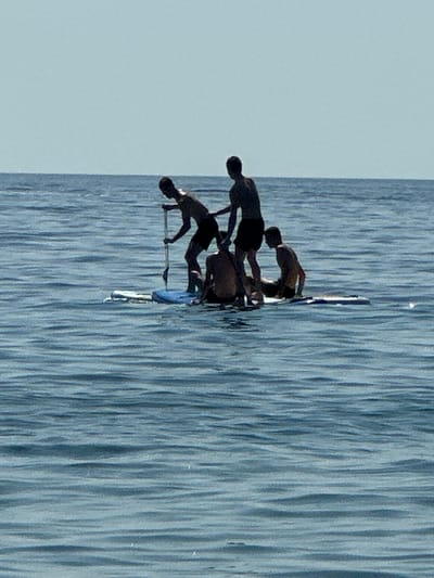 Four young males are on a paddleboard in the calm, blue sea under a clear sky. The paddleboard is partially submerged in the water, with a blue inflatable pad on top. One male is standing and holding a paddle, appearing to be paddling. Another male is standing behind him, and two other males are sitting on the paddleboard. The scene is outdoors, and the lighting suggests it is daytime. The location is identified as Đurovići, Montenegro.