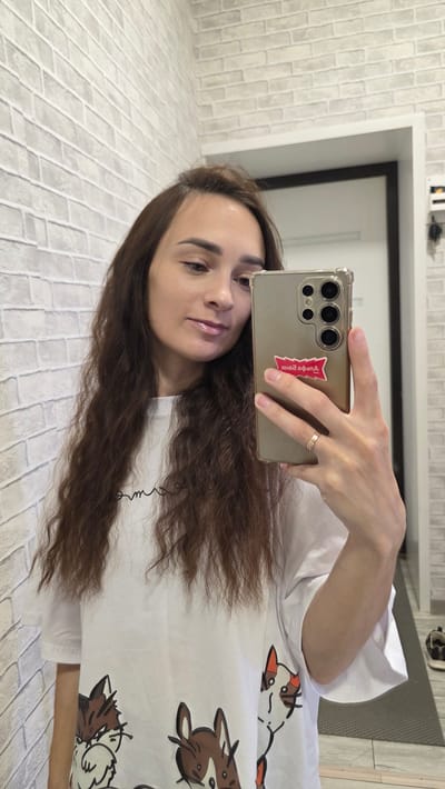 A young woman with long, wavy brown hair takes a selfie in a room with white brick-patterned walls. She is wearing a white t-shirt with cartoon cat illustrations on the front. Her right hand holds a gold-colored smartphone with a clear case, displaying a red sticker on the back. A ring is visible on her right ring finger. The background shows a doorway and a glimpse of a floor with a dark mat. The image appears to have been taken indoors.