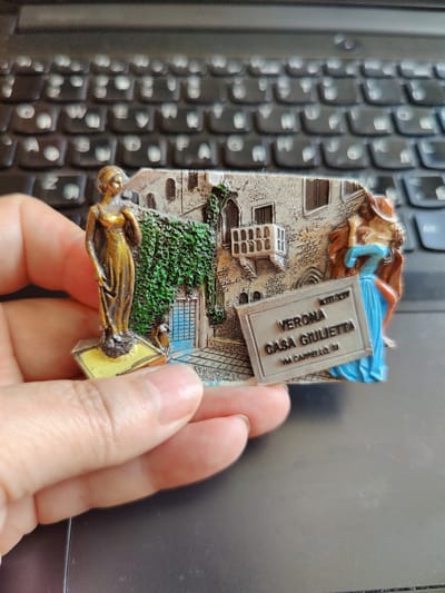 This media file features a close-up of a souvenir magnet held by a person's hand. The magnet depicts a miniature scene of Verona, Italy, specifically "Casa Giulietta" (Juliet's House). The scene includes two sculpted figures: a woman in a golden, flowing dress standing on a yellow base on the left, and another woman in a blue dress and brown cloak on the right, appearing to look upwards.
Behind the figures, there's a representation of a stone building with ivy growing on its facade, a balcony with railings, and a blue-paned window. A signpost with raised lettering is prominently displayed in the foreground.
Visible text on the signpost reads:
"XII-XIV"
"VERONA"
"CASA GIULIETTA"
"VIA CAPPELLO, 23"
The background of the image is a blurred view of a black keyboard. The lighting suggests an indoor setting, and there are no indicators of the time of day or weather. The focus is on the magnet and the hand holding it, highlighting the detailed craftsmanship of the souvenir. The context provided places this image as being taken in Rybnitsa, Moldova.