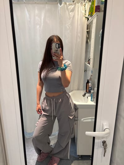 A young woman is taking a mirror selfie in what appears to be a bathroom. She is wearing a grey crop top and grey parachute pants with white stripes. She has long brown hair and is holding a smartphone in her right hand. On her left wrist, she is wearing a blue and green scrunchie. Her toenails are painted pink. She is wearing pink fuzzy slippers. The bathroom features a white shower curtain, a white sink with a faucet, and white cabinets. A white door with a white handle and keys is visible to the right. There are various toiletries on a shelf above the sink. The image is taken from a slightly low angle.