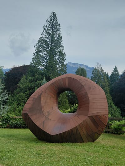 The image depicts a large, abstract, rusty metal sculpture in a grassy park. The sculpture is torus-shaped with a flattened opening, its surface is composed of many small, overlapping metal plates. In the background, a dense forest of various evergreen trees and a single large deciduous tree with dark red foliage are visible. Beyond the trees, a mountain range with a clear sky above is seen. The sculpture is situated on a well-maintained lawn with scattered white clover flowers. The setting is likely an outdoor park or garden in Levico Terme, Italy, as suggested by the natural landscape and the presence of sculptural art. There are no people or animals visible in the image.