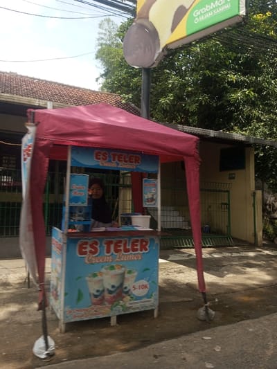 The image displays an outdoor street vendor stall in an Indonesian-speaking region, not Chori, Nigeria, as indicated by visible Indonesian text. A light-blue mobile food stall, featuring red lettering for "ES TELER Cream Lumes," illustrations of beverages, and a "5-10K" price range, is positioned prominently. It operates under a red canvas canopy supported by four poles weighted at their bases. Behind the counter, a person wearing a dark head covering and a light-colored top looks towards the viewer. Above and to the right, a sign for "GrabMart" with "SEJAM SAMPAI" (Arrives in an hour) in green text on a yellow background is mounted on a pole, alongside an illustration of a brown dessert. The background consists of a building with a brown tiled roof to the left, and a beige-walled building with a dark green metal gate and visible white steps to the right. Lush green foliage and overhead utility lines are present under a clear sky.