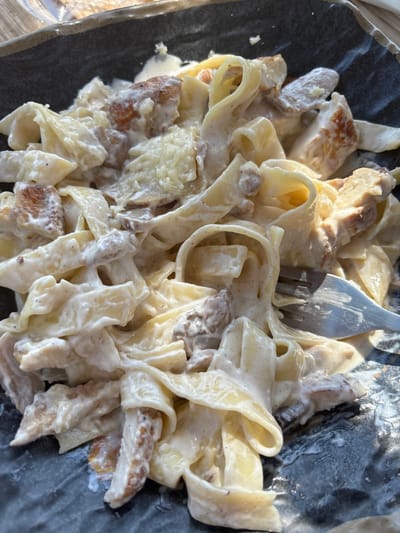 A close-up, overhead shot shows a plate of fettuccine Alfredo with chicken. The pasta is coated in a creamy white sauce, and pieces of grilled chicken are mixed throughout. A fork is visible on the right side of the plate, partially submerged in the pasta. The plate is dark gray and has a textured surface. The lighting suggests the photo was taken outdoors in daylight, as evidenced by the shadows and highlights. The background is blurred but appears to be a table. The setting is likely a restaurant in Al Yamānīyah, Jordan, given the context provided.
