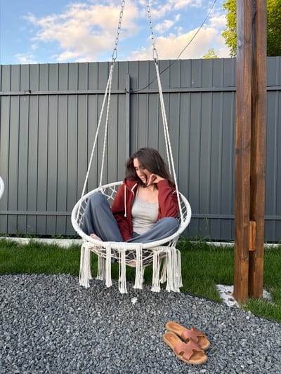 A young woman with brown hair sits in a white macrame swing chair on a sunny day in Sharovechka, Ukraine. She wears a rust-colored zip-up hoodie over a grey tank top and loose blue pants. Her legs are crossed, and her left hand rests on her chin as she looks down. The swing is suspended by a metal chain from a thick wooden post. Behind the swing is a dark grey corrugated metal fence, and to the left of the fence, the sky is visible with scattered clouds. The ground in front of the swing is covered with grey gravel, with a strip of green grass separating it from the fence. A pair of tan leather slide sandals lies on the gravel in the foreground.