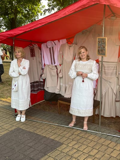 The image depicts an outdoor market stall in the city of Malaryta, Belarus. Two women are standing behind a stall covered by a red awning, displaying traditional Belarusian clothing, likely embroidered shirts and dresses. The woman on the left, with blonde hair, wears a long white dress with intricate embroidery on the chest and hem, and a small white crossbody bag. She has her arms crossed. The woman on the right, also with blonde hair, wears a white dress with similar embroidery on the bodice and hem. She is holding a small beaded item with a red tassel. Both women appear to be vendors. The stall itself is filled with various garments in neutral tones, with visible embroidery. The ground is paved with interlocking bricks in shades of brown and yellow. In the background, trees and other people are visible, suggesting a public gathering or event. The overall atmosphere is that of a local craft or cultural fair.