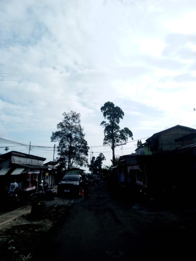 The image shows a street in Mya Sit Kaing Ywar Thit, Myanmar. The street is dark and appears to be unpaved. There is a car parked on the left side of the street, and several motorcycles are parked nearby. Two people are standing near the car and motorcycles. Buildings line both sides of the street. Two tall trees stand in the middle of the road. The sky is overcast. Electrical wires are visible above the buildings.