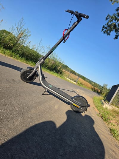 An electric scooter is parked on an asphalt path. The scooter is black with green lettering. A red cable lock is attached to the handlebars. The path is bordered by grass and vegetation. In the background, a fence and fields are visible under a clear blue sky. The scene is set in Godelheim, Germany.