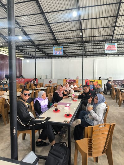 The image depicts an indoor dining area, likely a food court or restaurant, in Panteraya, Indonesia. A group of approximately ten people, appearing to be friends or family, are seated at a long rectangular table, engaged in conversation and enjoying food and drinks. The group is diverse in age and gender, with several women wearing headscarves.

On the left side of the image, a man wearing sunglasses and a black jacket is smiling at the camera. Next to him is a young girl in a purple puffer vest. Further down the table, several individuals are interacting, with some looking at their phones and others gesturing. A woman in the foreground on the right is holding a drink and making a peace sign with her fingers.

The environment is characterized by an open ceiling with exposed metal beams and corrugated roofing, illuminated by fluorescent lights. Several signs are visible on the ceiling, including one in blue that reads "DILARANG BERMAIN" (No Playing) with associated icons and another in red that states "ABRISAM COFFEE KAMI TIDAK MEMFASILITASI SEGALA BENTUK KEMAKSIATAN JUDI, NARKOBA DAN PROSTITUSI" (ABRISAM COFFEE WE DO NOT FACILITATE ANY FORM OF IMMORALITY, GAMBLING, DRUGS AND PROSTITUTION).

The table is set with drinks in glasses and red saucers, along with what appear to be small food items. Brown plastic chairs surround the table. In the background, other tables and chairs are visible, along with signage for "DIMSUM" and "Okky T". The floor is concrete. The overall atmosphere appears casual and social.