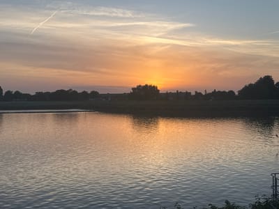 The image shows a sunset scene over a body of water, likely located in Capelle aan den IJssel, Netherlands, given the flat landscape characteristic of the region. The sky is filled with warm hues of orange and yellow transitioning to cooler tones of blue and grey. A single contrail cuts across the upper portion of the sky.

The water reflects the colors of the sunset. The landscape in the midground appears as a dark silhouette with trees and buildings. The foreground shows the edge of the water with some vegetation along the bank. A metal railing is also visible in the lower right corner. There are no visible people.