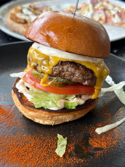 A top-down, close-up shot captures a burger on a dark plate, along with a blurred dish of food in the background. The burger appears to be cut in half, with the top bun slightly separated and held in place by a metal skewer. The burger consists of a beef patty, melted yellow cheese, white sauce, tomato slices, pickles, and lettuce. Orange powder-like seasoning is scattered on the plate around the burger. In the background, the blurred dish shows what appears to be a pizza with toppings that include red onions. Based on the prompt, the location is in Salaspils, Latvia.