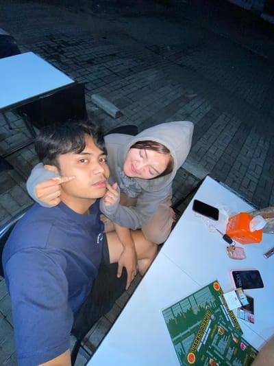 The image captures a close-up view of a young couple at an outdoor seating area, likely a cafe or restaurant, in Mekar Bakti, Indonesia. The lighting suggests it is evening or night.

In the foreground, a young man is seated, wearing a navy blue t-shirt. He is looking directly at the camera, with a slightly pensive expression. A young woman, wearing a grey hooded sweatshirt and tan shorts, is leaning in from behind him. Her left hand is gently touching his left cheek, while her right hand forms a heart shape near his face. She is smiling and looking down at him.

On a white table in front of them, several items are visible. A black smartphone is placed near the top of the table. Next to it is an orange tissue box with a white tissue peeking out. A set of car keys with a pink keychain is also on the table, along with a pink smartphone. Two green menus, titled "WARKOP AGAM 99", are spread out on the table, displaying lists of food and drinks. The background consists of a paved ground with interlocking bricks, and what appears to be outdoor furniture, including a table with a light blue surface.