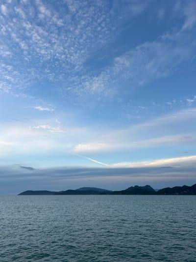 The image displays a serene natural landscape with a clear blue sky filled with wispy clouds. A thin contrail cuts diagonally across the sky, hinting at distant air traffic. In the background, a silhouetted mountain range stretches across the horizon. The foreground is dominated by the calm, rippling surface of the ocean, its color a muted blue-green. The lighting suggests either early morning or late afternoon, with soft illumination and long shadows cast by the mountains. The overall mood is peaceful and tranquil. The scene appears to be a coastal view, possibly from a boat or a vantage point overlooking the sea and mountains. There is no visible text in the image.