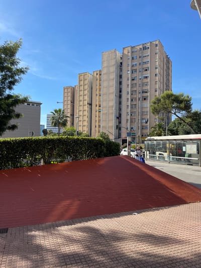 The image depicts an urban scene in Cornellà de Llobregat, Spain, characterized by apartment buildings, a bus stop, and a park area. In the foreground, a red, sloped surface, possibly a decorative element or part of a park structure, is visible, covered in tile on the right side. Beyond this, a meticulously trimmed green hedge forms a boundary. To the right, a bus stop with a transparent enclosure shows people inside, and a bus is partially visible. The background is dominated by several tall apartment buildings with a repetitive facade structure, typical of mid-20th-century urban architecture. A palm tree and other greenery suggest a Mediterranean climate. A man is seen walking with a blue bag next to the bus stop. The sky is a clear, bright blue with a few wispy clouds. The overall impression is that of a sunny day in a residential urban area.