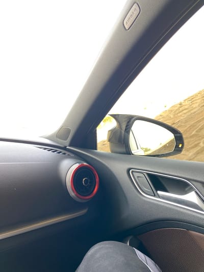 The image shows the interior of a car, likely an Audi, with a focus on the passenger side door and dashboard. The car is parked in a desert-like environment, characterized by sandy hills in the background. The dashboard features a circular air vent with a red and silver accent. The side mirror is visible, reflecting the exterior landscape. The "AIRBAG" label is prominently displayed on the A-pillar. The overall scene suggests a road trip or a stop in a remote, arid location. The specific location in Boughezoul, Algeria, cannot be determined from the image alone, as it lacks any identifying landmarks or signs specific to that city.