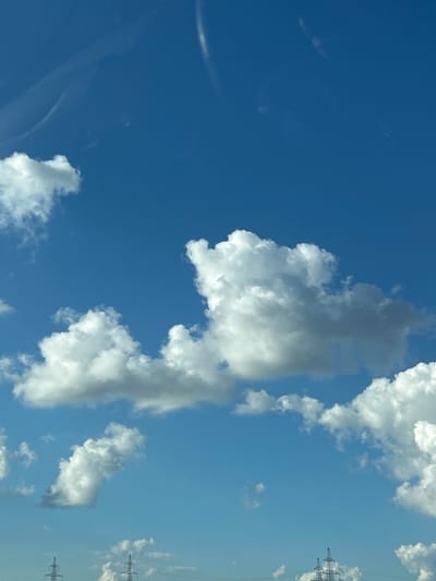 The image captures a clear blue sky with scattered cumulus clouds. In the lower portion of the frame, the tops of several electricity pylons are visible, suggesting an outdoor, possibly semi-rural or industrial, setting. The lighting indicates it is daytime. There are no people or animals depicted in the image. The composition focuses on the sky and the distant infrastructure. The scene does not provide any specific visual cues to definitively place it in Primorskii, Russia, beyond the provided metadata context.