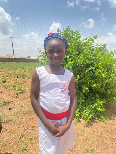 A young girl in a white dress with a red sash and a blue cap stands outside in Turu, Nigeria. She is smiling and looking towards the camera. Behind her is a green bush, and in the background, there is a field with low-lying plants. The ground is brown and dry, with patches of green grass. The sky is blue with scattered white clouds. Electrical lines are visible in the distance on the left. A brick wall and corrugated iron roof can be seen beyond the field.