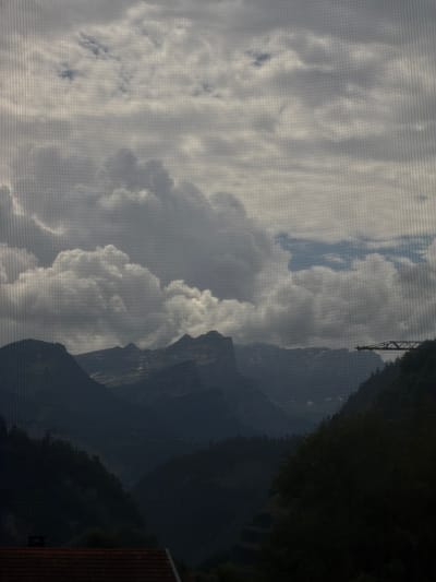 The image depicts a mountainous landscape viewed through a screen, likely a window screen, which creates a faint grid pattern across the scene. The sky is filled with dramatic, billowing clouds, ranging from bright white to dark gray, suggesting an overcast or stormy day. Below the clouds, silhouetted mountains rise, their dark forms contrasting with the lighter sky. The peaks are jagged and appear to be covered in snow in some areas. The lower foreground shows a dark, forested hillside and a portion of a reddish-brown tiled roof, indicating the presence of human habitation. To the right, a construction crane is visible against the sky, its skeletal structure outlined. The overall mood is somber and dramatic, with a sense of the power of nature. The specific location within Cluses, France is not discernible from the image itself; the image depicts a generalized mountainous scene.