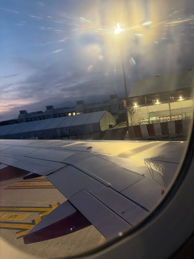 The image, taken from an airplane window, shows an airport scene at dusk. The sky is a dusky blue with hints of pink and orange near the horizon, suggesting either sunrise or sunset. A bright light source, possibly a runway or tarmac lamp, is visible in the upper left, creating lens flare effects with streaks of light radiating outwards.

In the foreground, the wing of the airplane is prominently featured, extending from the right side of the frame. The wing is a light gray color, and its surface appears slightly wet, with streaks of water visible. There are markings on the wing, including the number "2".

Adjacent to the wing, the airport terminal building is visible. It is a modern structure with large glass windows that reflect the lights from inside and outside. The building appears to be multi-story, with several sections. There are also visible air conditioning units mounted on the exterior of some parts of the building. Yellow painted lines and markings, typical of an airport tarmac, are visible beneath the wing. One marking appears to be "E175".

The scene depicts an airplane parked at an airport gate, likely preparing for departure or having just arrived. The wet tarmac and the dimming light suggest a potentially damp evening or early morning. No people are clearly visible, and the overall mood is calm and functional, characteristic of an airport environment. The location is identified as Lamezia Terme, Italy, but this information is not visually discernible from the image itself.