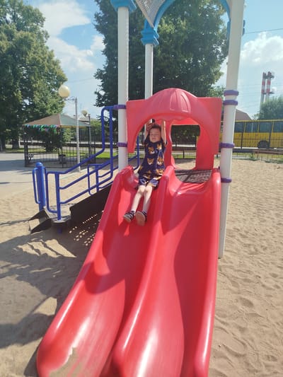 A young boy is sitting on a red plastic slide on a sunny day in a playground located in the city of Yalutorovsk, Russia. The boy is wearing a dark blue shirt with a tiger print and shorts, and is looking at the camera with a slight smile. He is positioned on the upper part of the slide, with his legs extended down. The slide is part of a larger playground structure with blue railings and white poles. The ground beneath the slide is covered in sand, and there are some shadows cast on the sand. In the background, there are trees, a fence, a gazebo, and a yellow bus. There is also a tall industrial chimney with red and white stripes visible in the distance. The sky is blue with some white clouds.