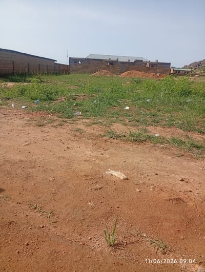 This is a medium-angle, eye-level, full, landscape photograph of a dirt and grass area in DU, Nigeria, on June 11, 2026, at 9:04 AM. In the background, there is a brick structure that appears to be under construction, with visible rebar and unfinished walls. A pile of red dirt is also present. To the left, a partially constructed brick wall stretches into the distance. The foreground is dominated by a dirt path with scattered tufts of grass and some litter, including plastic bottles. The sky is clear and bright.