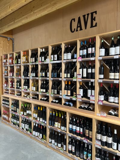 This image captures a well-stocked wine cellar or a dedicated wine section of a store, seen from a low, upward-looking perspective and rotated counter-clockwise. Hundreds of wine bottles are neatly organized in extensive wooden shelving units, which create individual compartments for each bottle. The shelving is constructed from light-colored wood, contrasting with a darker wooden wall on the left and a gray, concrete-like wall on the right side of the background.
The shelves display a wide variety of wines, including red, white, and rosé, distinguished by the bottle shapes, label designs, and the liquid's color. Many bottles feature intricate labels, with some text partially visible, such as "Steph.", "BALAN DE BALARD", "LINGUART", "Chant d'Assians", and "CHATEAU". Small white and colored tags (pink, green, purple) are attached to the shelves, likely indicating prices or product information.
A prominent, dark-colored sign spelling "CAVE" (French for "cellar" or "wine cellar") is affixed to the wooden wall on the left, reinforcing the setting as a wine storage or sales area. The wines appear meticulously arranged, possibly by type, region, or producer, making for an organized display. The scene is brightly lit, suggesting indoor artificial lighting. No people are visible. Given the location context of Loire-Authion, France, the scene depicts a typical French wine retail environment, offering a diverse selection of wines for purchase.