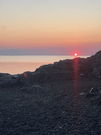 This is a photograph of a sunset over a body of water, likely Lake Kapchagay, in the city of Karashygan, Kazakhstan. The sun, a bright red orb, is low on the horizon, casting a warm glow across the sky and water. The foreground is dominated by a rocky, uneven shoreline covered in dark stones and pebbles. Jagged, dark grey rocks rise in the mid-ground, creating a rugged texture. The water is calm and reflects the soft colors of the sunset, with subtle ripples visible. In the distance, a hazy landmass or island can be seen along the horizon line. The overall atmosphere is serene and peaceful, characteristic of a sunset over a large body of water.