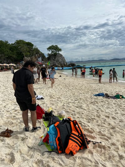 In Tambon Phru Nai, Thailand, on a crowded beach with white sand, a group of people are visible. The sky is overcast with gray clouds. In the foreground, a man in a black short-sleeved shirt and black shorts stands facing away from the camera, looking down. Near his feet, brown sandals and a pile of beach gear, including orange and black life vests, bags, and a blue inflatable toy, are scattered on the sand. Further down the beach, individuals are gathered near the turquoise water, some wading in or standing by inflatable flotation devices. In the background, rocky outcrops with vegetation rise from the sea, and a line of blue floating barriers separates the swimming area. Several colorful umbrellas are set up on the sand further inland.