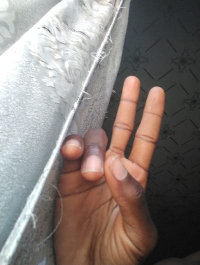 A dark-skinned human left hand is positioned prominently in the foreground, making a 'V' gesture with the index and middle fingers extended upwards. The thumb, ring, and pinky fingers are folded. Finger creases and clean fingernails are visible. To the upper left, a light-colored fabric, possibly grey or silver with a textured, subtly patterned (likely floral or abstract swirl) surface, extends vertically, revealing a hemline with visible loose threads. The fabric suggests a curtain or upholstery. In the background, to the right of the fabric and behind the hand, a darker surface exhibits a repeating geometric pattern of tessellated squares or diamonds, each containing a four-petal motif. This background element suggests an interior wall or floor. Lighting appears to originate from the left, illuminating the hand and fabric more brightly than the background. The scene is located within an interior space in Kaninkon, Nigeria.